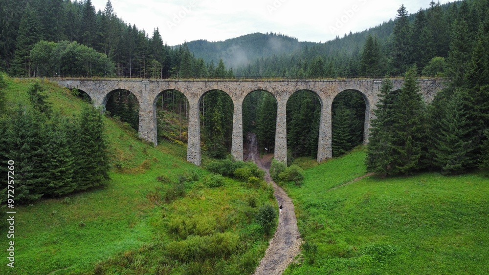 Fototapeta premium A stunning drone video of Chmarošský Viaduct in Slovakia showcases its impressive stone arches, nestled in lush forests and scenic mountain landscapes