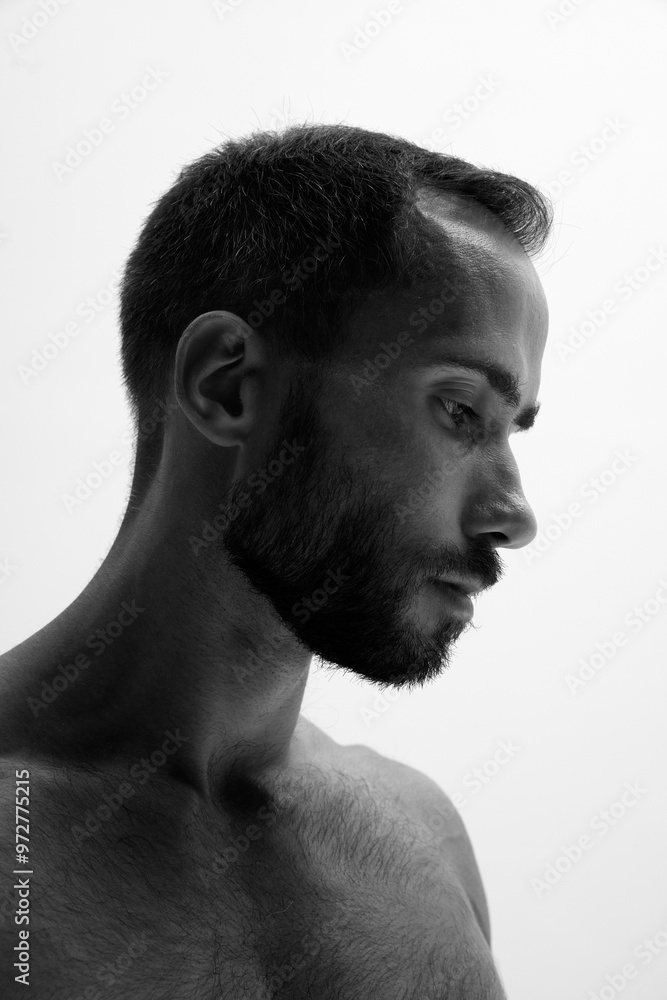 Bearded man stands in profile with thoughtful expression against white background. Photo in black and white filter. Concept of beauty and fashion, style and youth, male health. Bearded man stands in profile with thoughtful expression against white background. Photo in black and white filter. Concept of beauty and fashion, style and youth, male health.