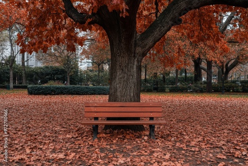 A wooden bench sits under a large tree in a park, surrounded by a blanket of vibrant red and orange fall leaves, capturing the essence of autumn in a serene outdoor setting.