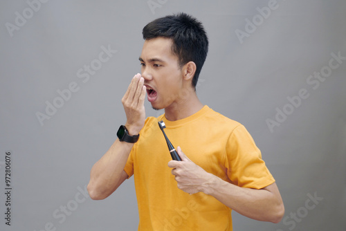 A young Asi man in a yellow t-shirt stands on a gray background, holding an electric toothbrush. He covered his mouth with his hand and looked disgusted by the problem of bad breath photo