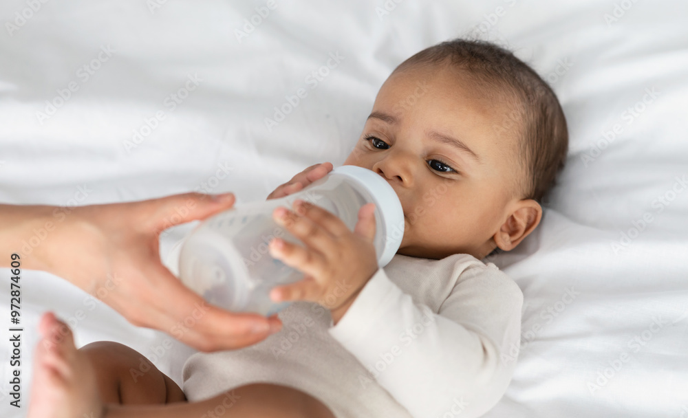 Childcare And Nutrition Concept. Portrait of cute little African American baby drinking milk from bottle, woman helping and holding, kid wearing bodysuit lying on the white blanket at home Childcare And Nutrition Concept. Portrait of cute little African American baby drinking milk from bottle, woman helping and holding, kid wearing bodysuit lying on the white blanket at home