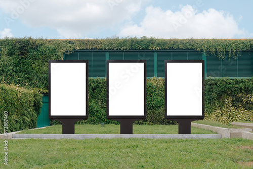 Vertical mock up template: 3 out of home (OOH) advertising billboard posters on grass, against background of lush plant wall of building. Bright day, blue sky with clouds. Transparent png file.
