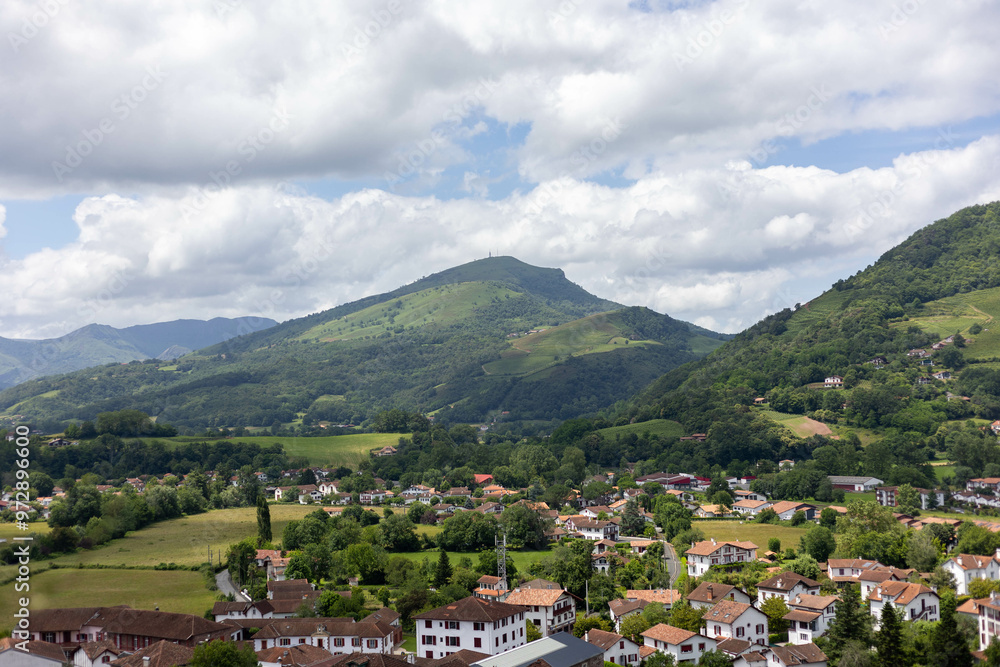Fototapeta premium Paysage du Pays basque Français avec un ciel nuageux, une montagne et un petit village entouré de forêt et de champs vert