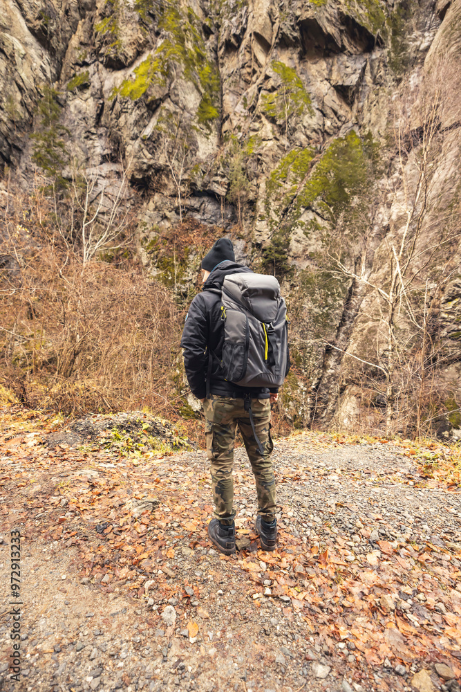 Amidst golden foliage and towering cliffs, a lone adventurer stands on a trail, captivated by nature's beauty while carrying a sturdy backpack for exploration Amidst golden foliage and towering cliffs, a lone adventurer stands on a trail, captivated by nature's beauty while carrying a sturdy backpack for exploration