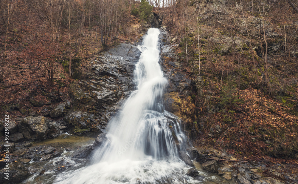 Obraz premium Rushing water tumbles gracefully over rugged stones, while surrounding trees display vibrant autumn colors, creating a serene natural escape away from urban life