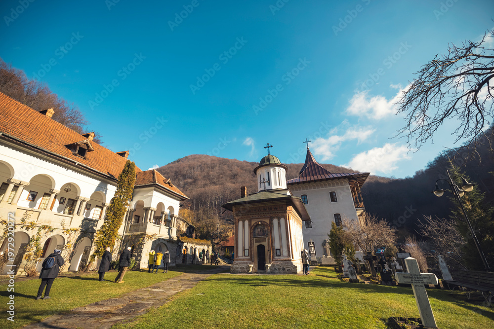 Naklejka premium Visitors stroll through the lush grounds of a historic monastery framed by majestic mountains, basking in the warm sunlight of a late afternoon