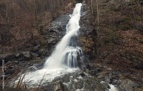 A powerful waterfall tumbles down rugged stones, creating a silken flow of water. Surrounding trees stand tall, while the early light filters through the foliage, adding a magical aura
