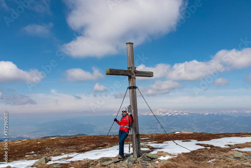 Hiker woman reaching wooden summit cross standing atop mountain peak Lenzmoarkogel, Gleinalpe, Lavanttal Alps, Styria, Austria. Hiking trail on snow covered alpine meadow. Wanderlust. Tranquil serene photo