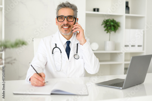 The grey-haired doctor exhibits a confident demeanor, speaking on the phone while looking at the laptop, indicating a modern approach to patient consultations and healthcare technology.