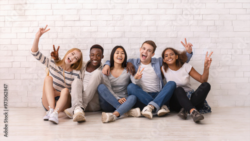 Emotional multiracial students sitting on floor, showing peace gesture, posing at camera, white wall background