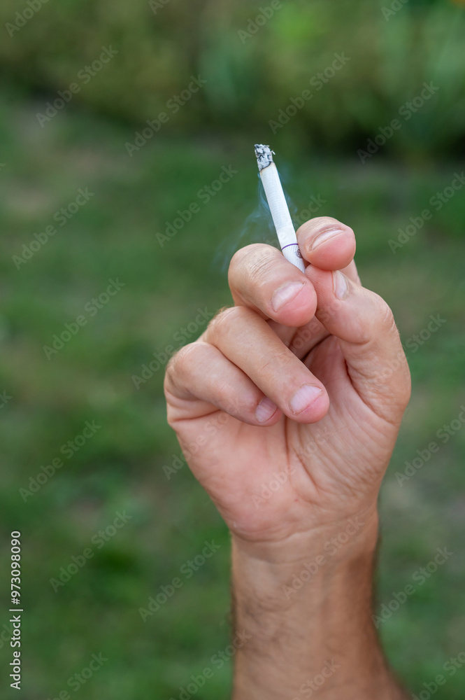 A man's hand holds a cigarette in his hand against a background of green grass. A man's hand holds a cigarette in his hand against a background of green grass.