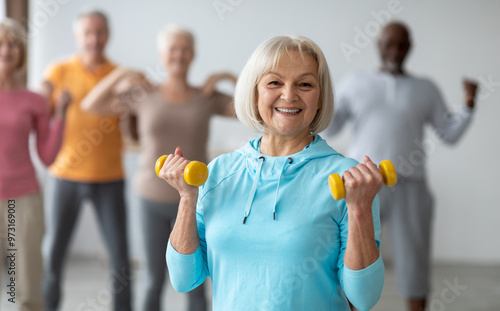 Fototapeta Naklejka Na Ścianę i Meble -  Multiracial group of senior people in sportswear doing strength building fitness exercises with dumbbells, holding fitness tools and smiling at camera, selective focus on positive elderly lady