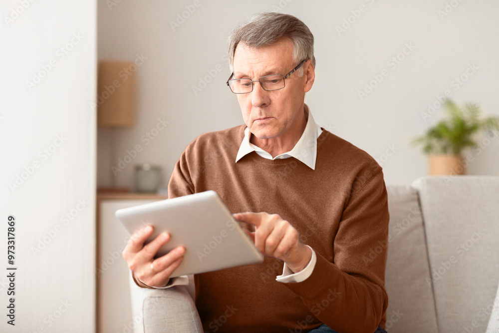 Serious Elderly Man Using Digital Tablet Browsing Internet And Reading Business News Sitting On Couch Indoor. Selective Focus Serious Elderly Man Using Digital Tablet Browsing Internet And Reading Business News Sitting On Couch Indoor. Selective Focus