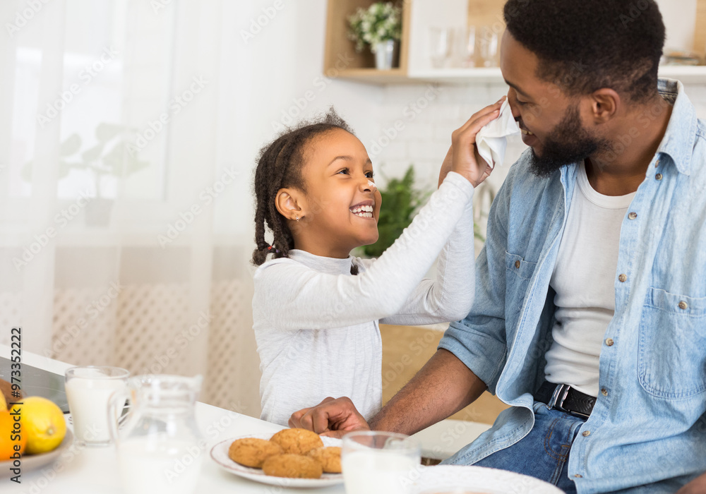 Afro Man And His Daughter Having Fun At Breakfast, girl cleaning father nouse with tissue, kitchen interior, copy space Afro Man And His Daughter Having Fun At Breakfast, girl cleaning father nouse with tissue, kitchen interior, copy space