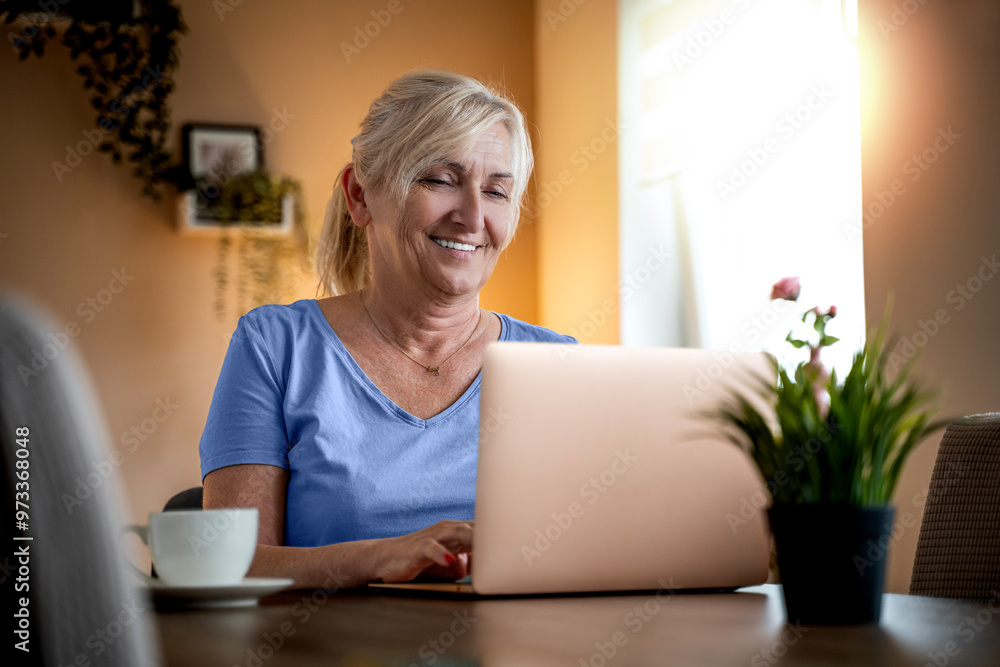 Smiling middle aged senior woman working at home sitting at table in living room. Happy mature woman using laptop technologies and writing. Blonde with eyeglasses work indoors Smiling middle aged senior woman working at home sitting at table in living room. Happy mature woman using laptop technologies and writing. Blonde with eyeglasses work indoors