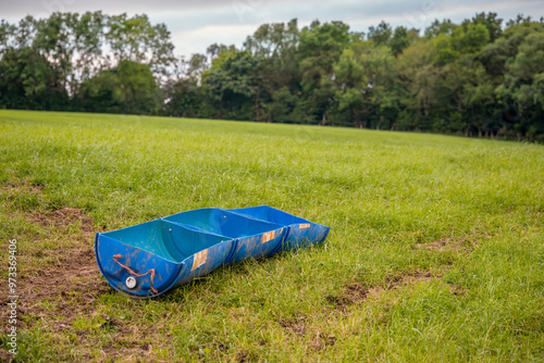 Farm field cattle animal feeding trough homemade from split 220 litre blue plastic barrel on grass slope with trees and hedge