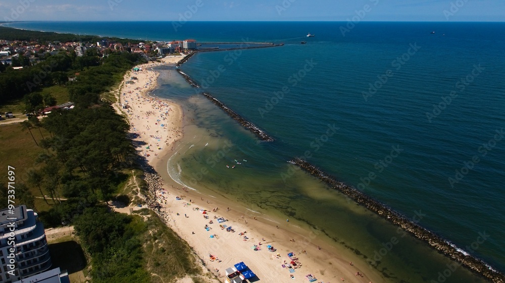 Obraz premium Drone captures Darłowo’s golden beach, sunbathers, swimmers, and ships heading out of the port into the calm Baltic Sea.