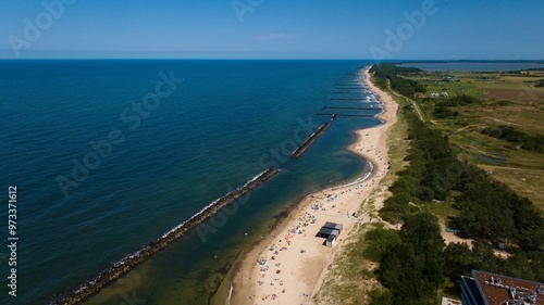 Aerial view of Darłowo beach, with golden sand, calm Baltic Sea, beachgoers, hotels, and dunes with forest.