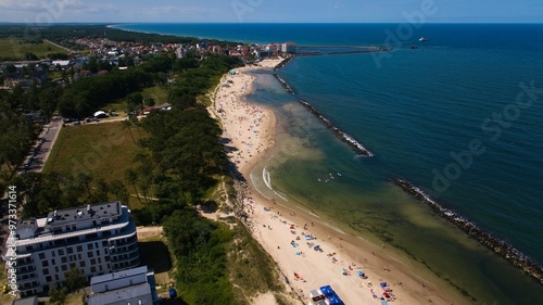 Aerial shot of Darłowo beach, showing golden sand, calm waters, beachgoers, and ships sailing from the port.