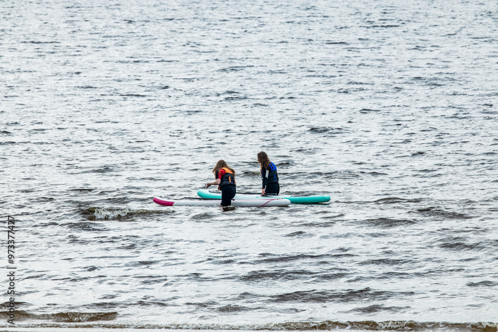 Obraz premium Two female paddle boarders on boards in sea water with island with lighthouse and hills on horizon