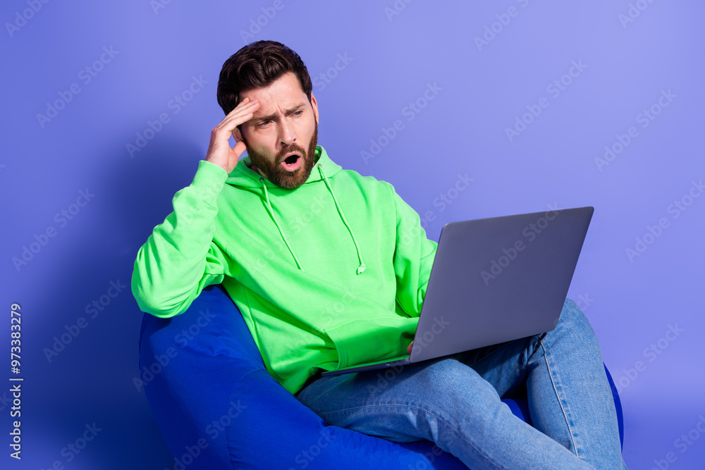 Photo of handsome young guy sit beanbag hold netbook nervous worried dressed stylish green garment isolated on violet color background Photo of handsome young guy sit beanbag hold netbook nervous worried dressed stylish green garment isolated on violet color background