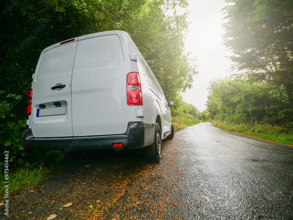 A white medium size van is parked on a small county road in rural area with a red tail light. The road is empty and the sun is shining brightly. Product delivery to remote locations concept.