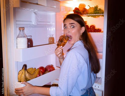 Midnight snack. Scared girl in pajamas eating croissant near opened fridge, free space