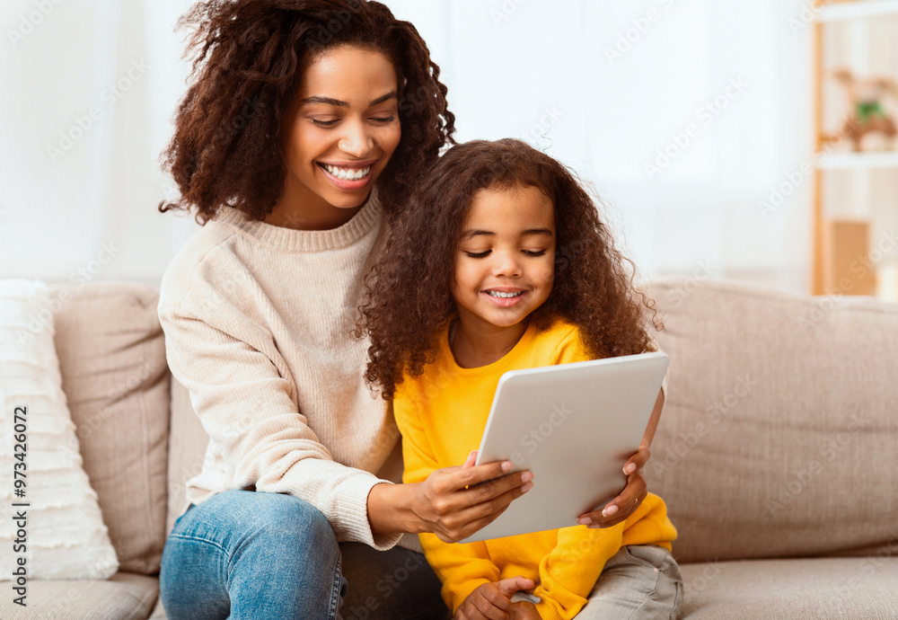 African American Mother And Daughter Using Digital Tablet Computer Together Sitting On Sofa At Home. Weekend Family Leisure African American Mother And Daughter Using Digital Tablet Computer Together Sitting On Sofa At Home. Weekend Family Leisure