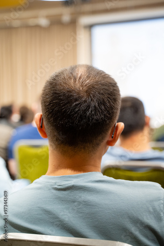 A man who is wearing ear buds can be seen sitting in a lecture hall