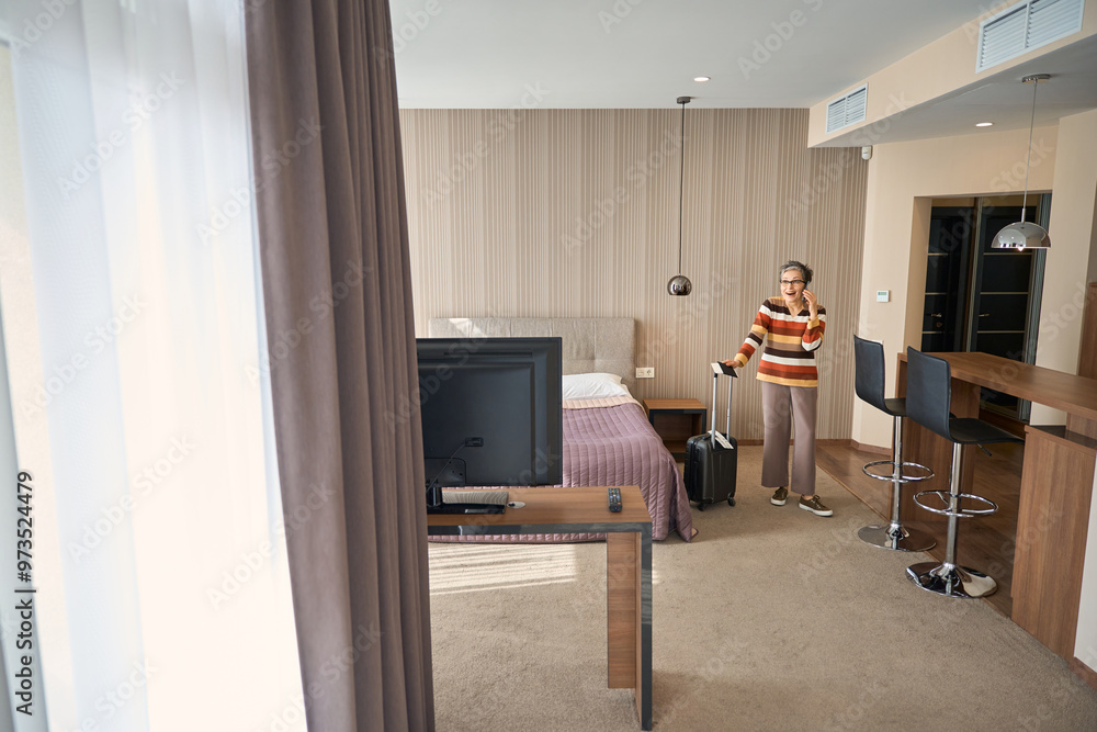 Happy adult woman with baggage, travel documents talking on phone in hotel room Happy adult woman with baggage, travel documents talking on phone in hotel room