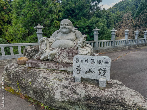 A joyful stone statue of the Laughing Buddha seated on a large rock, surrounded by greenery, with a traditional Japanese lantern and sign, symbolizing peace and happiness in a serene setting.