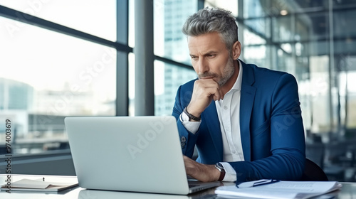 A handsome middle-aged businessman is sitting at his desk in the office, looking pensive and deeply focused on working with a laptop computer while holding his hand under his chin.