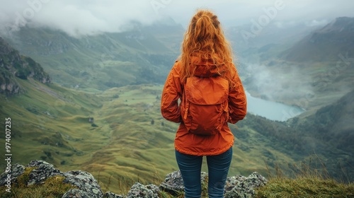 A young woman in an orange jacket gazes at a misty mountain valley while standing on a rocky outcrop during a cloudy day
