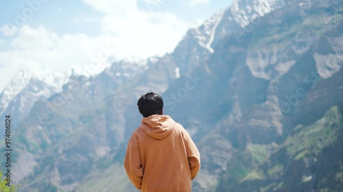 4K Rear view shot of an Indian male tourist wearing hoodie walking in front of snowy Himalayan mountains at Marbal village in Lahaul Valley, Himachal Pradesh, India. Tourist in front of Himalayas. photo