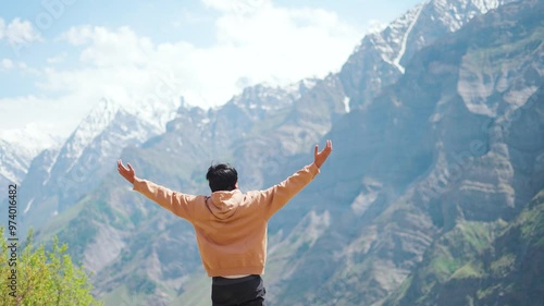 4K Rear view shot of an Indian male tourist wearing warm hoodie raising his arms in front of the snowy Himalayan mountain peaks at Marbal village in Lahaul Valley, Himachal Pradesh, India.  photo