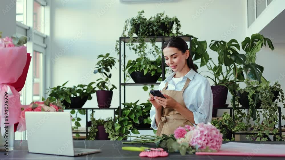 Young, successful female florist receiving an order on her mobile phone. She smiles and selects a few pink carnations and begins to arrange a bouquet. Young, successful female florist receiving an order on her mobile phone. She smiles and selects a few pink carnations and begins to arrange a bouquet.