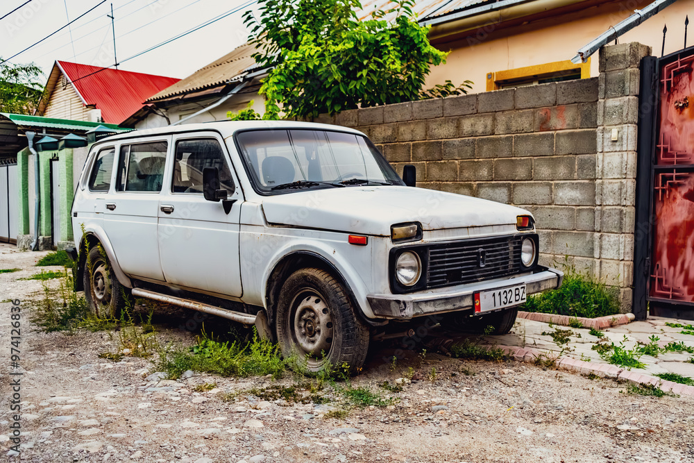 Fototapeta premium A vintage white Soviet-era Lada parked on a quiet, tree-lined residential street in Bishkek, Kyrgyzstan. The weathered car stands as a nostalgic reminder of Soviet history, blending seamlessly with th