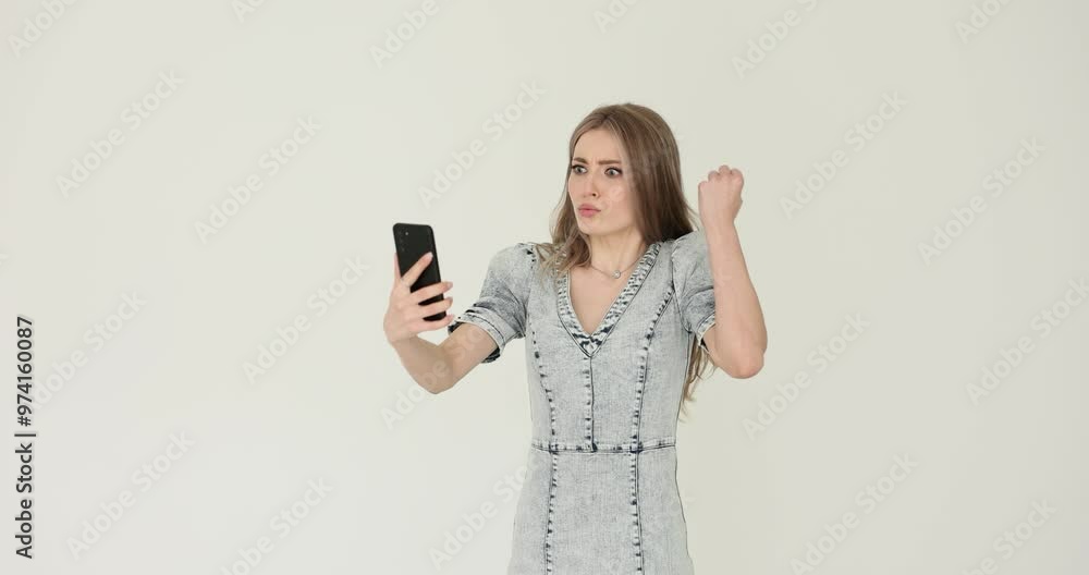 Emotional young woman expresses irritation during video call against white. Female expressively demonstrates negative feelings holding smartphone Emotional young woman expresses irritation during video call against white. Female expressively demonstrates negative feelings holding smartphone