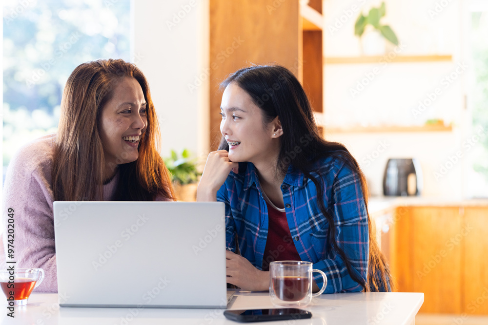 At home, Smiling asian mother and daughter using laptop and drinking tea in kitchen At home, Smiling asian mother and daughter using laptop and drinking tea in kitchen