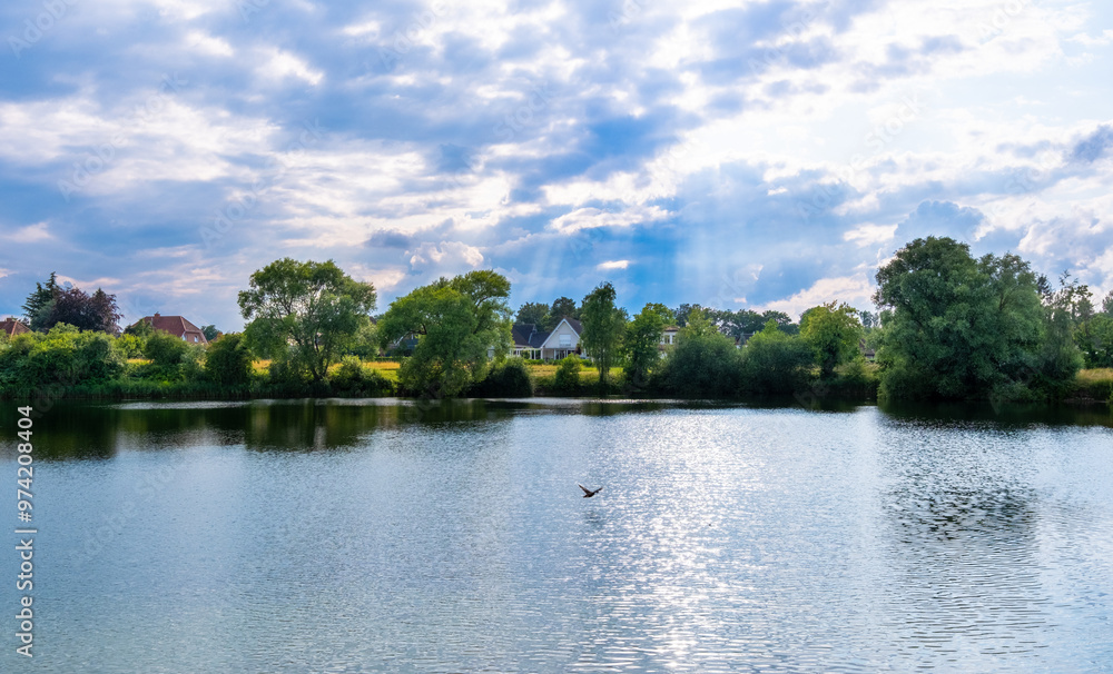 Peaceful lakeside view with trees, houses, and soft sunlight breaking through the clouds Peaceful lakeside view with trees, houses, and soft sunlight breaking through the clouds