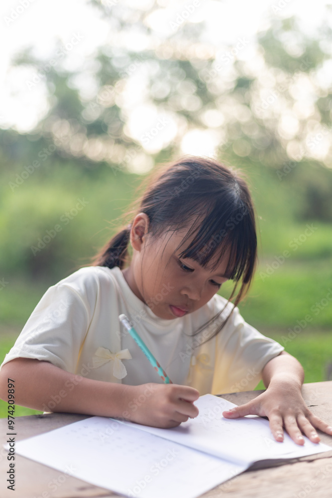 A young girl is sitting at a table writing. She is wearing a white shirt and has her hair in a ponytail. A young girl is sitting at a table writing. She is wearing a white shirt and has her hair in a ponytail.