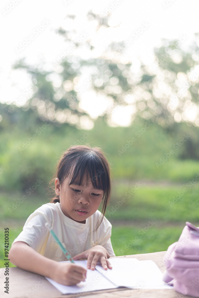 A young girl is sitting at a table writing. She is wearing a white shirt and has her hair in a ponytail. A young girl is sitting at a table writing. She is wearing a white shirt and has her hair in a ponytail.