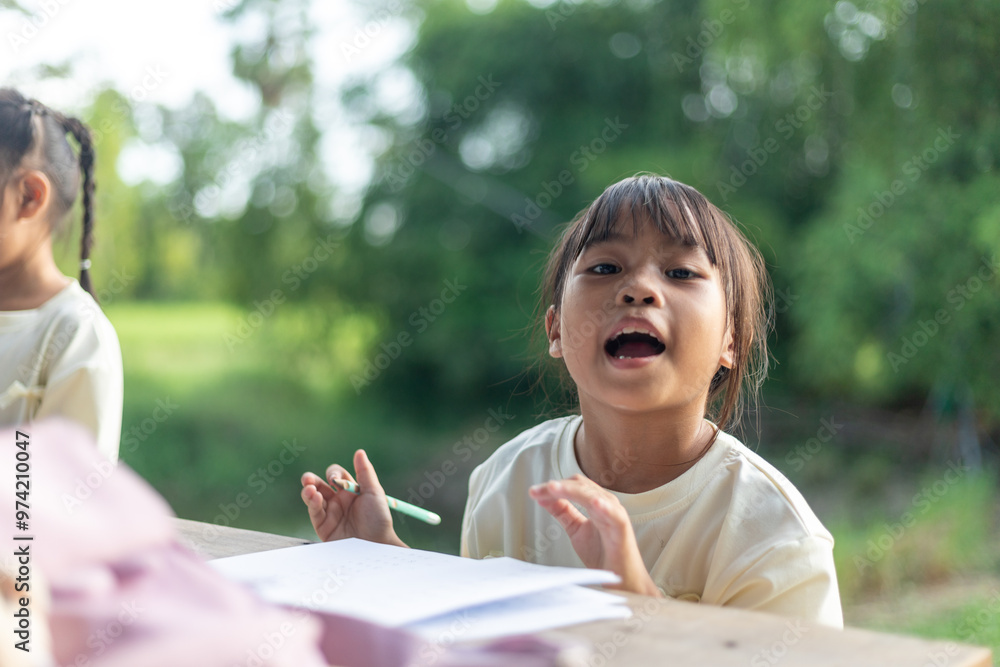 A young girl is sitting at a table writing. She is wearing a white shirt and has her hair in a ponytail. A young girl is sitting at a table writing. She is wearing a white shirt and has her hair in a ponytail.