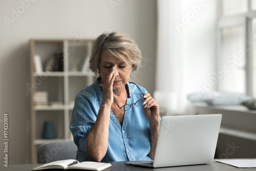 Photos Mature woman takes off eyeglasses sit at desk with laptop, experiences discomfort or tiredness, due to eyestrain, remove glasses, rubbing nose bridge