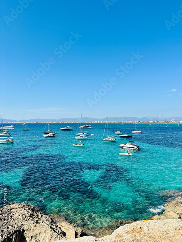 View from the rocky shore to the bay with yachts in the azure sea and with mountains and a city on the horizon