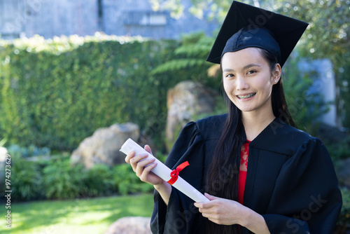 In garden, Graduating asian young woman holding diploma and smiling outdoors in cap and gown photo