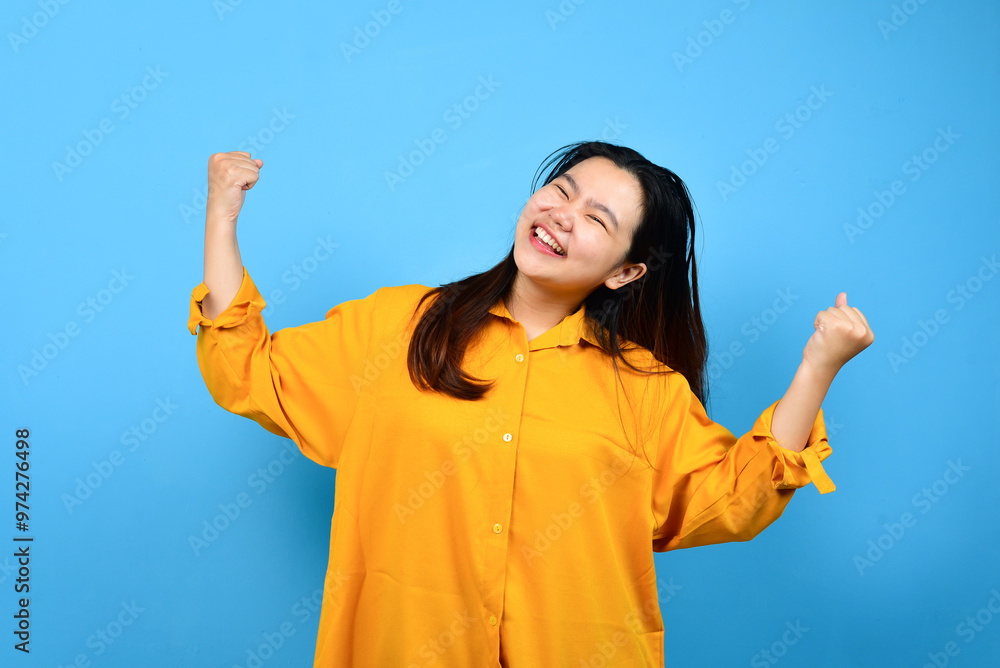 A young Asian woman with a happy successful expression wearing yellow shirt with copy space isolated on blue backround A young Asian woman with a happy successful expression wearing yellow shirt with copy space isolated on blue backround