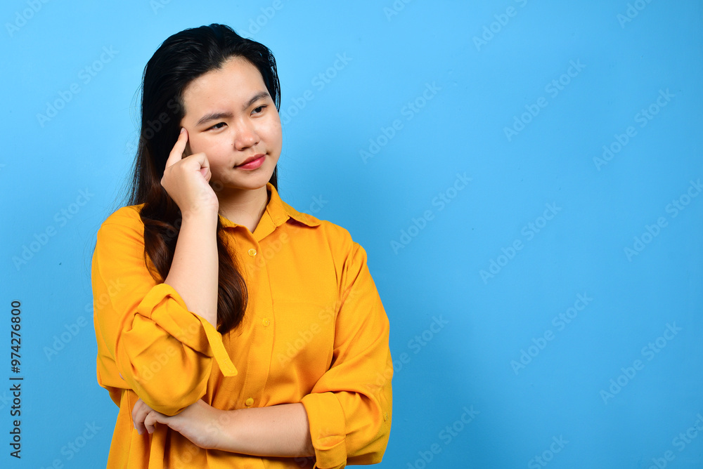 A confused young woman wearing a yellow shirt while looking to the side, isolated on a blue background with copy space A confused young woman wearing a yellow shirt while looking to the side, isolated on a blue background with copy space