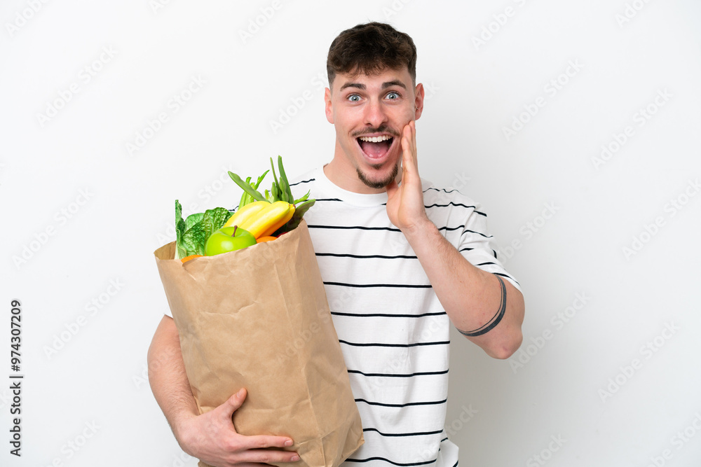 Young caucasian man holding a grocery shopping bag isolated on white background with surprise and shocked facial expression Young caucasian man holding a grocery shopping bag isolated on white background with surprise and shocked facial expression