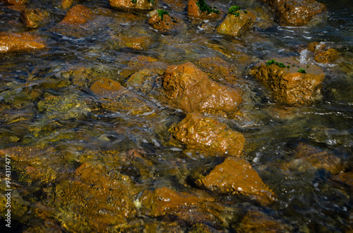 a rocky coastline with vibrant orange-brown rocks partially covered in green moss with gentle waves create frothy white foam as they wash over the stones